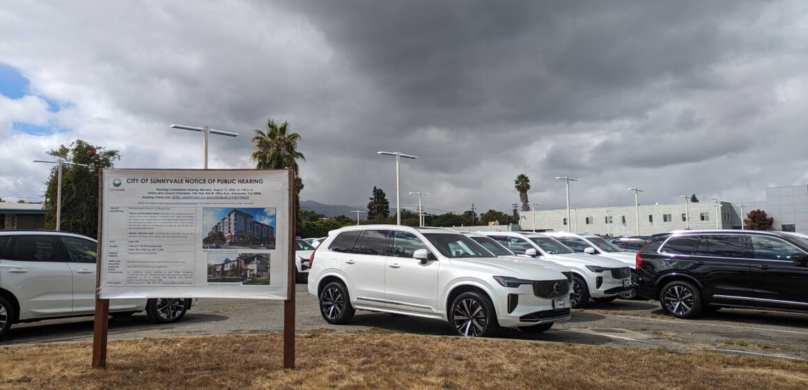 A public hearing notice sign for a development stands in front of cars parked at a dealership