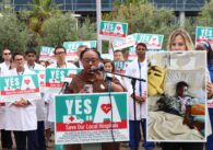A woman speaks at a podium with a crowd of people behind her holding signs