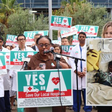 A woman speaks at a podium with a crowd of people behind her holding signs