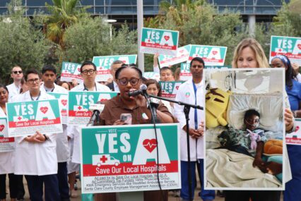 A woman speaks at a podium with a crowd of people behind her holding signs