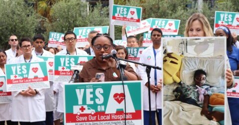 A woman speaks at a podium with a crowd of people behind her holding signs