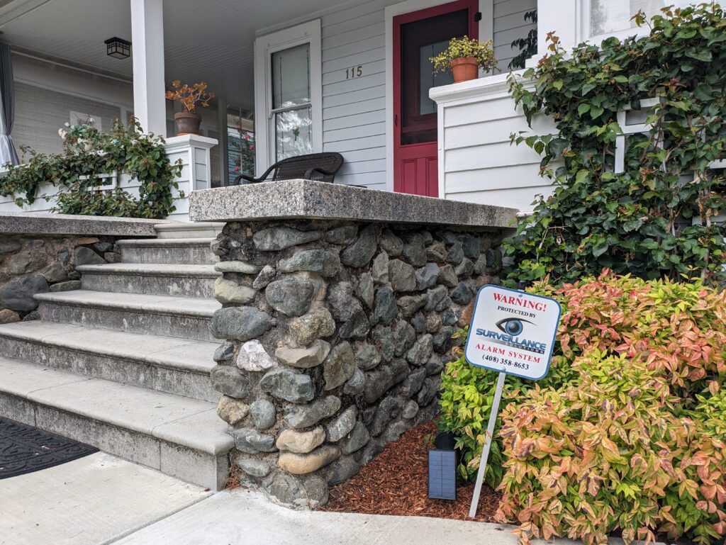 A sign sticks out of the ground in a resident's front yard that reads, "Warning! Protected by Surveillance Solutions alarm system."