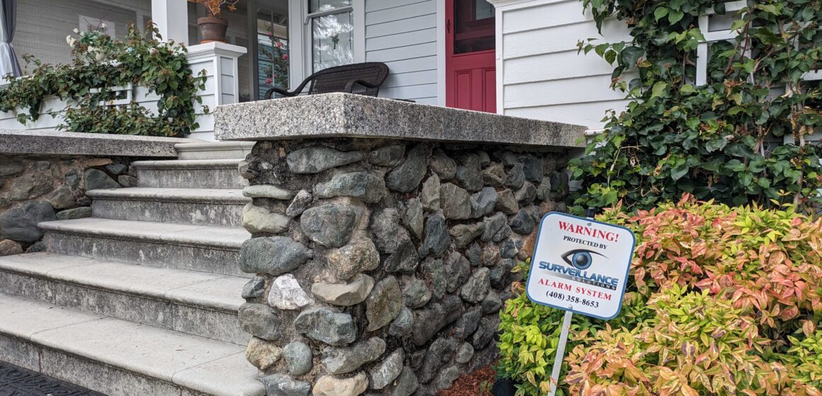 A sign sticks out of the ground in a resident's front yard that reads, "Warning! Protected by Surveillance Solutions alarm system."