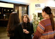 A woman facing the camera speaks to two other women with their backs to the camera