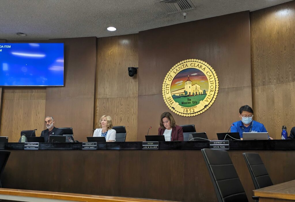Two women and two men sit behind a dais in front of a sign that reads "The City of Santa Clara California"