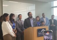 A man speaks at a podium, surrounded by other people