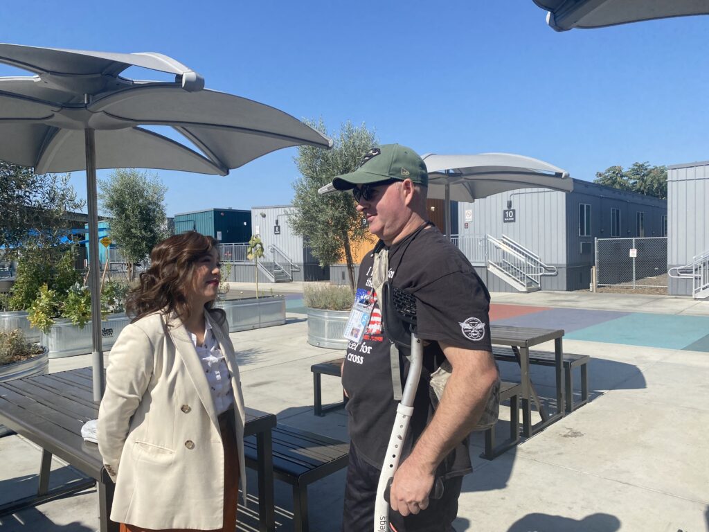 A woman in a coat outside speaks to a man in a t-shirt and baseball hat using crutches