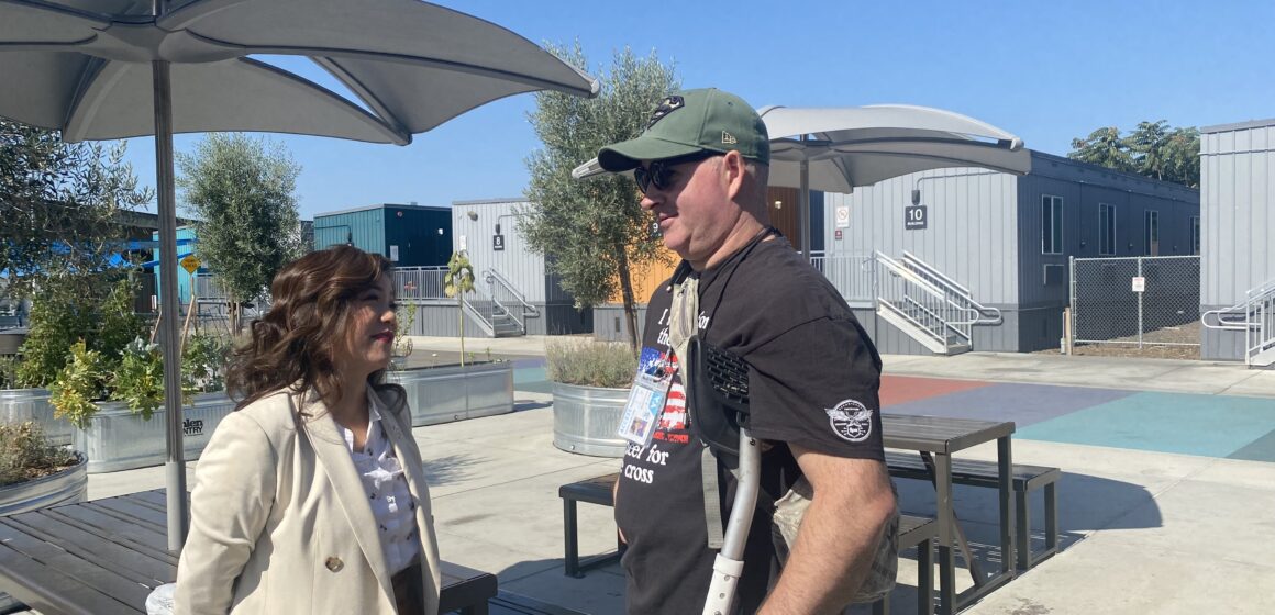 A woman in a coat outside speaks to a man in a t-shirt and baseball hat using crutches