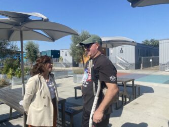 A woman in a coat outside speaks to a man in a t-shirt and baseball hat using crutches