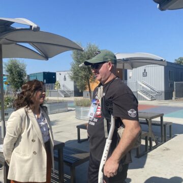 A woman in a coat outside speaks to a man in a t-shirt and baseball hat using crutches