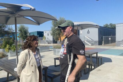 A woman in a coat outside speaks to a man in a t-shirt and baseball hat using crutches
