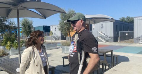 A woman in a coat outside speaks to a man in a t-shirt and baseball hat using crutches