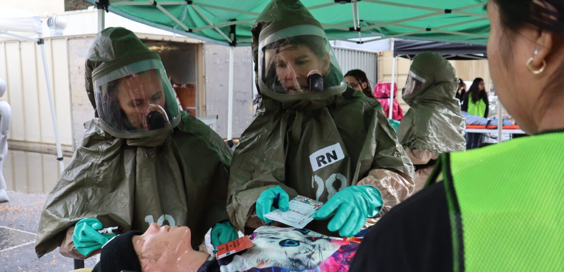 Two people in hazmat suits practice evaluating a medical mannequin