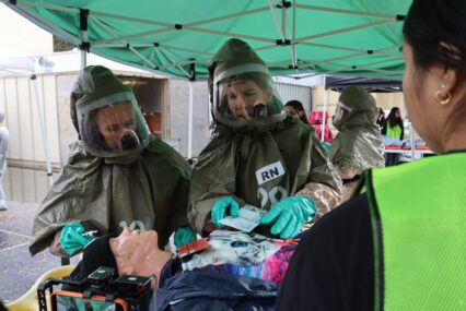 Two people in hazmat suits practice evaluating a medical mannequin