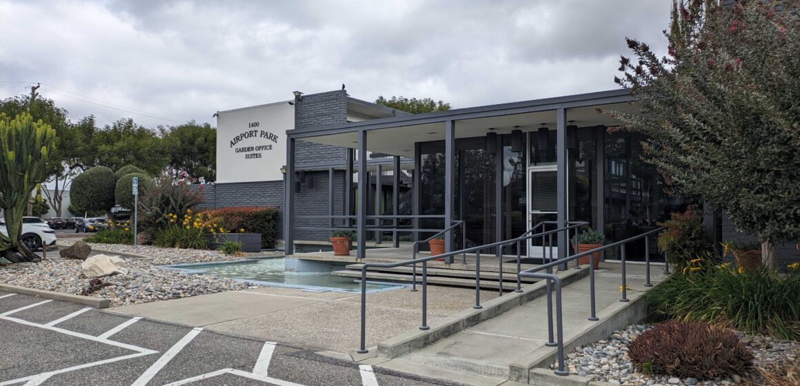 The front of a grey and white building with a sign that reads "Airport Park Garden Office Suites"