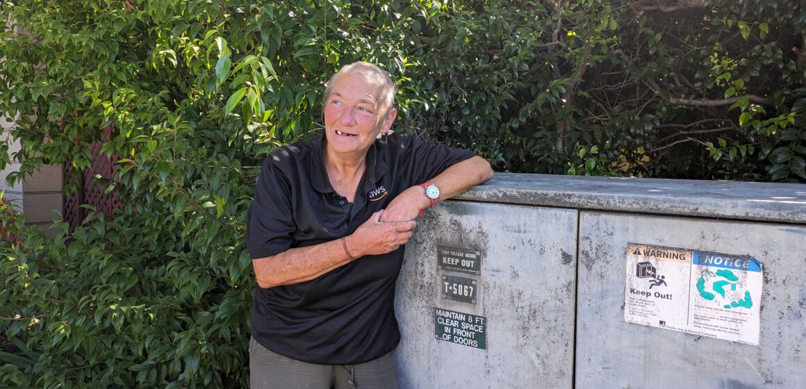 An older homeless woman with a close-shaved buzz cut leans on a utility box