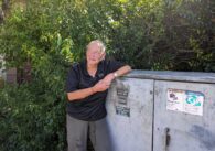 An older homeless woman with a close-shaved buzz cut leans on a utility box