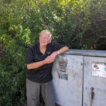 An older homeless woman with a close-shaved buzz cut leans on a utility box