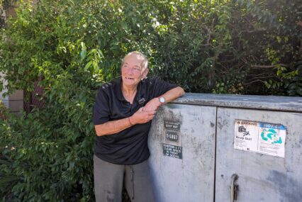 An older homeless woman with a close-shaved buzz cut leans on a utility box