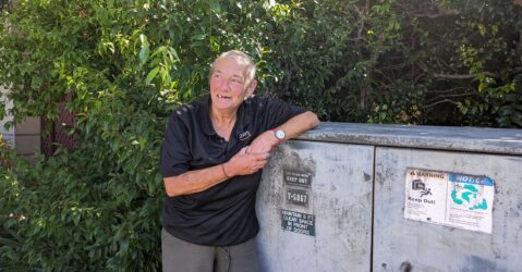 An older homeless woman with a close-shaved buzz cut leans on a utility box