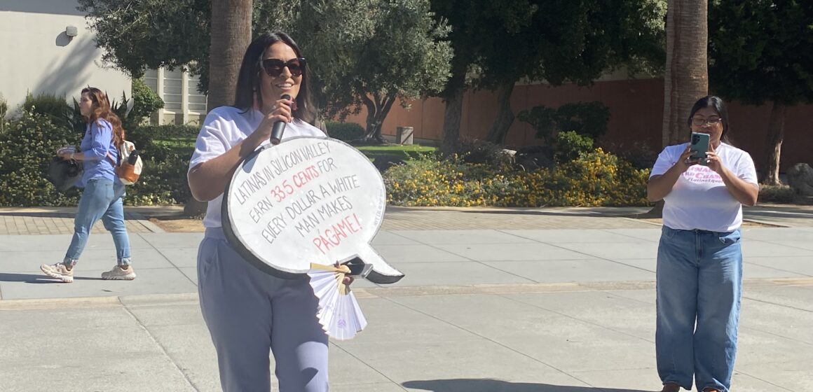 LATINA EQUAL PAY DAY A woman stands outside holding a sign