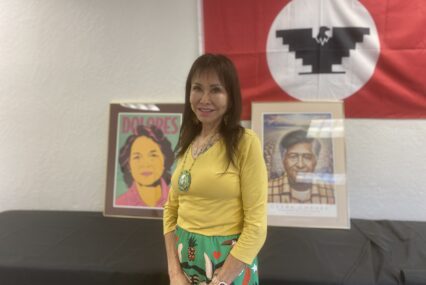 A woman stands in front of a United Farm Workers flag and art depicting civil rights leaders Dolores Huerta and Cesar Chavez.