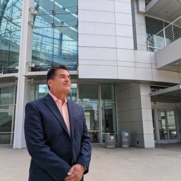 Một người đàn ông lớn tuổi mặc áo khoác vest nhìn vào khoảng không trước một tòa nhà tròn lớn màu bạc