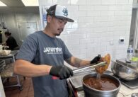 A man in a restaurant kitchen wearing black gloves uses tongs to dip chicken into a pot of hot oil