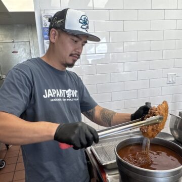 Scratch Cookery A man in a restaurant kitchen wearing black gloves uses tongs to dip chicken into a pot of hot oil