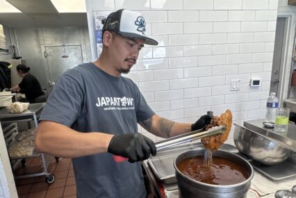 A man in a restaurant kitchen wearing black gloves uses tongs to dip chicken into a pot of hot oil