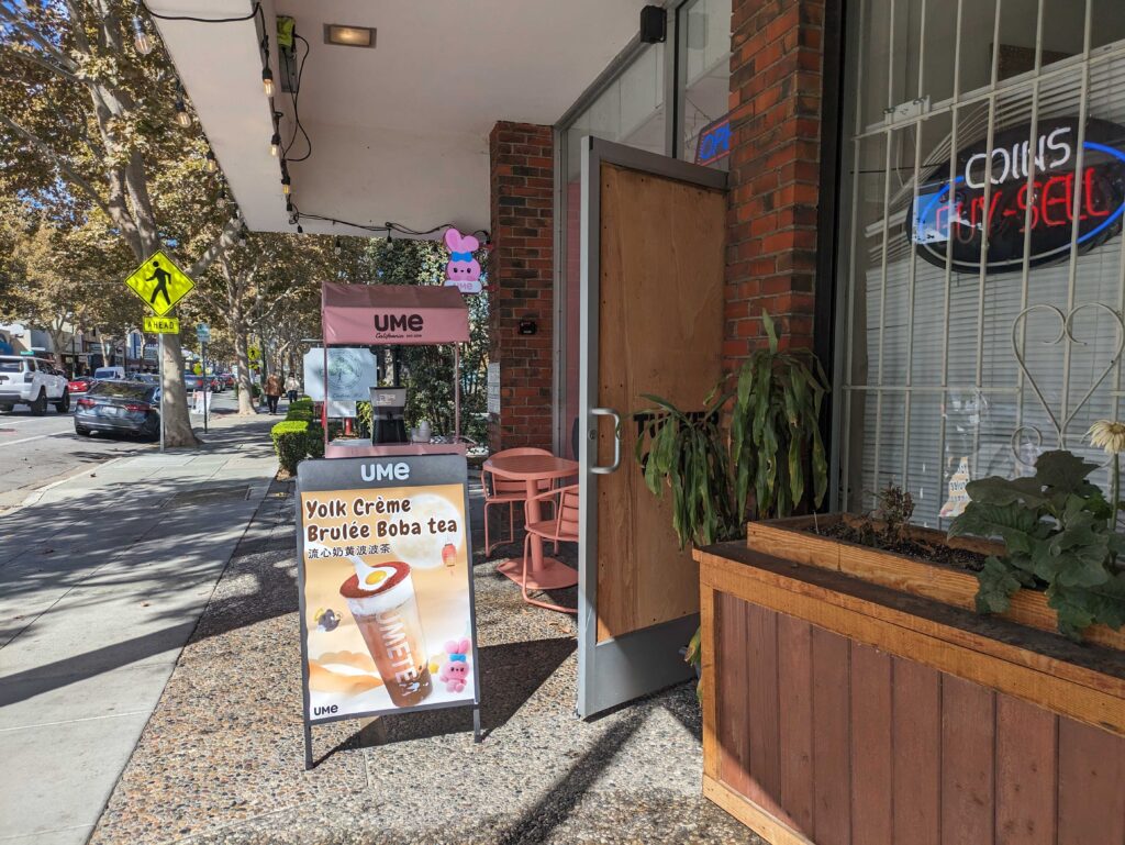 A shop door where the glass has been replaced by wood, next to an a-frame sign that reads "Yolk Creme Brulee Boba Tea"