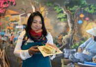 A woman in an apron and scarf holds a plate of food inside a restaurant