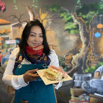 A woman in an apron and scarf holds a plate of food inside a restaurant
