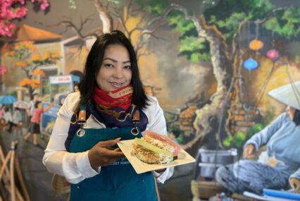A woman in an apron and scarf holds a plate of food inside a restaurant