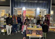 A group of children and adults stand in front of West Valley Community Services surrounded by groceries