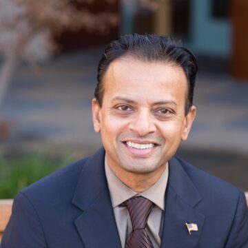 Rishi Kumar Un hombre con traje y un pin con la bandera estadounidense sentado en un banco