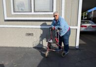 An older woman with short gray hair stands in front of her mobile home, leaning on a walker