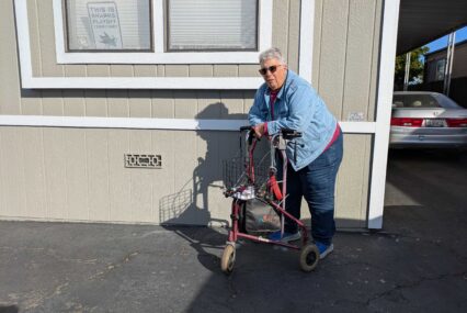 An older woman with short gray hair stands in front of her mobile home, leaning on a walker
