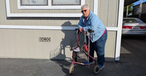 An older woman with short gray hair stands in front of her mobile home, leaning on a walker