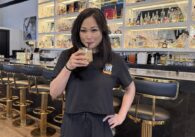 A woman sips a drink from a straw in a restaurant, with a bar behind her
