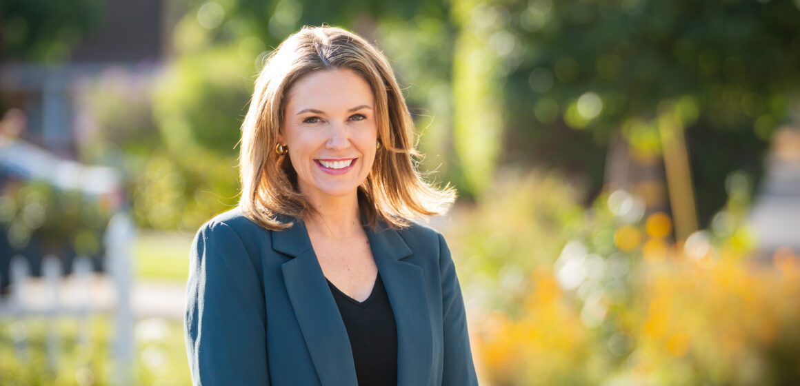 A woman in a turquoise suit jacket and jeans stands in front of a city street.