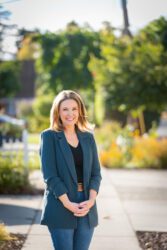 Genny Altwer A woman in a turquoise suit jacket and jeans stands in front of a city street.