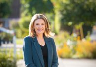 A woman in a turquoise suit jacket and jeans stands in front of a city street.
