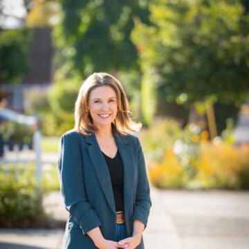 Genny Altwer A woman in a turquoise suit jacket and jeans stands in front of a city street.