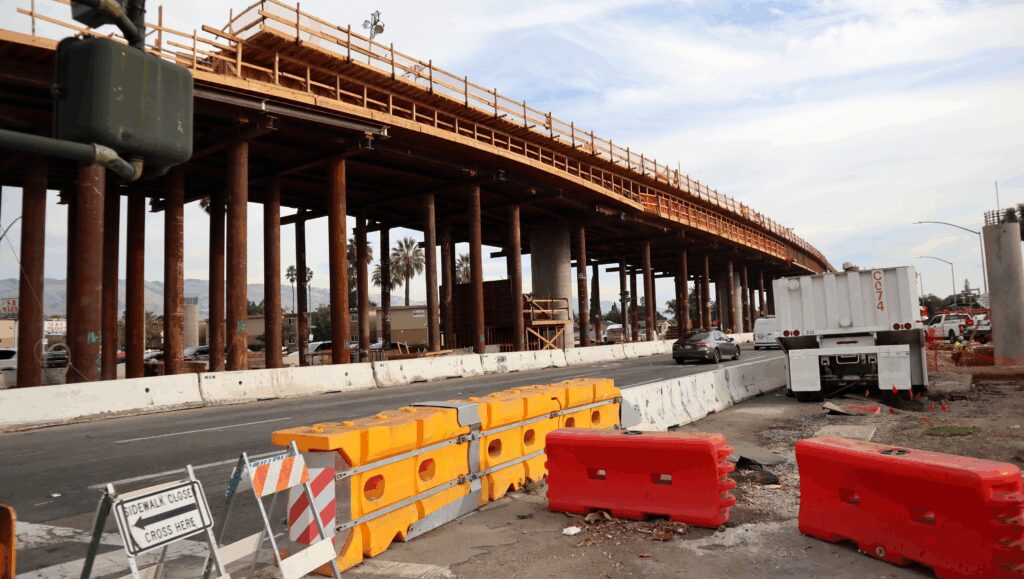 Photo of a three lane road with massive construction on both sides, closing off the sidewalk on one side and a raised platform under construction on the other