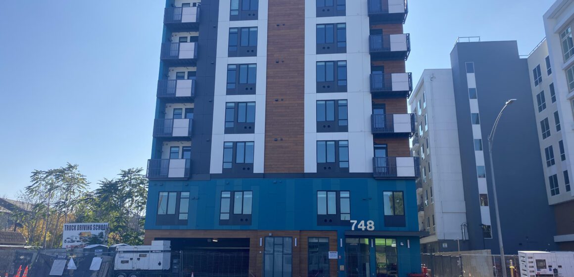 Un edificio residencial de siete pisos con detalles en blanco, naranja y gris que se eleva sobre un sitio en construcción.