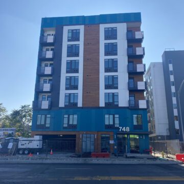 A seven-story residential building with white, orange and gray trim rising up from a construction site.