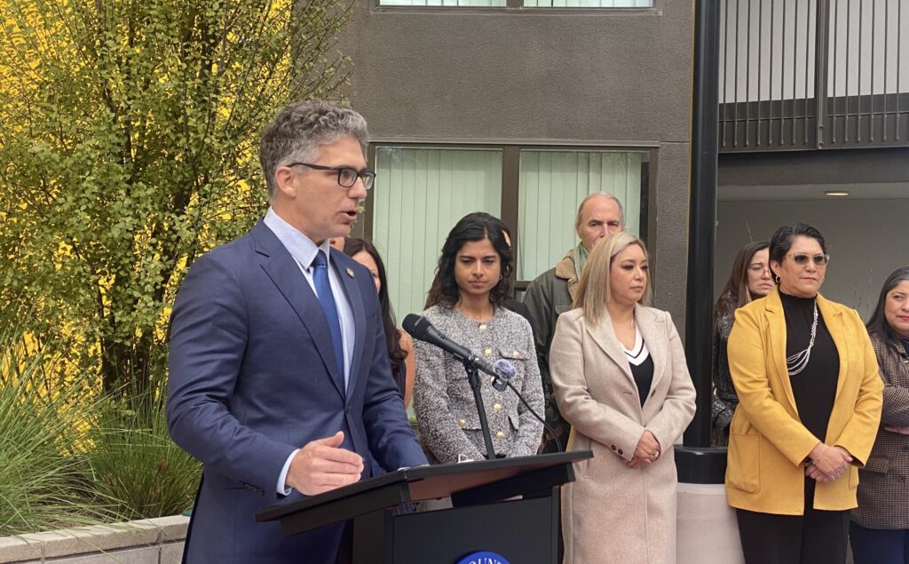 Một người đàn ông mặc vest và thắt cà vạt nói vào micro trên bục phát biểu, với những người đứng phía sau anh ta