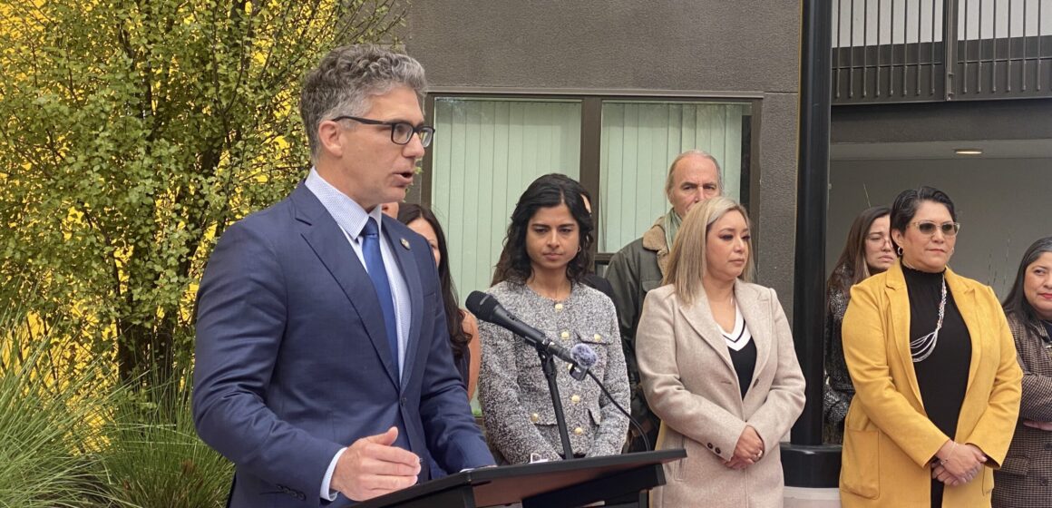 A man in a suit and tie speaks into a microphone at a podium, with people standing behind him