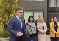 A man in a suit and tie speaks into a microphone at a podium, with people standing behind him